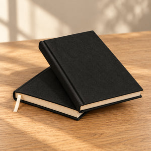 Two blank black canvas notebooks stacked on a wood table in natural light | Pamusan.com