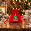 Bolsita para regalos de terciopelo rojo, diseño ¡Feliz Navidad! con acebo, en una mesa rústica nevada ante el árbol
