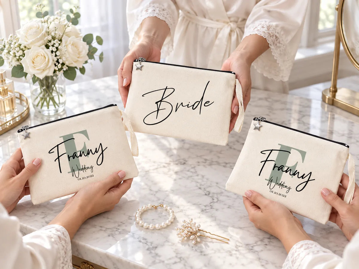 Manos de damas de honor con batas de seda sobre un tocador de mármol, sosteniendo un clutch Bride y dos clutches Franny Wedding de algodón, con rosas blancas y una pulsera de perlas
