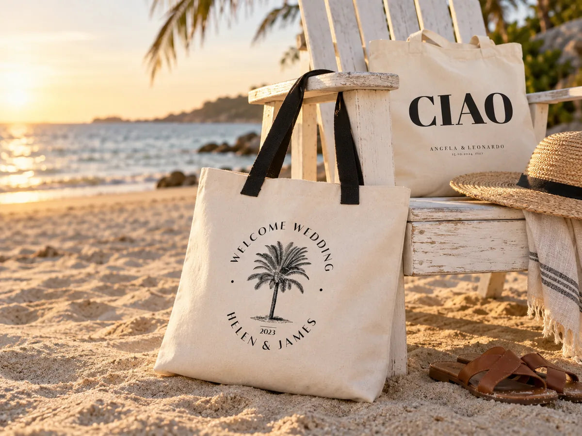 Gran bolsa de lona Welcome Wedding Helen & James con palmera y asas negras apoyada en una silla Adirondack blanca en la playa a la hora dorada, con una bolsa CIAO, un sombrero de paja y sandalias de cuero detrás